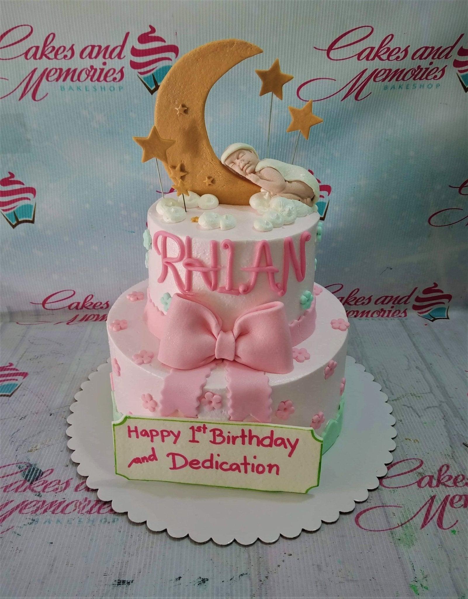 Pink 2-tier baby shower cake with a sleeping baby on a crescent moon topper, pink bow, and floral accents.