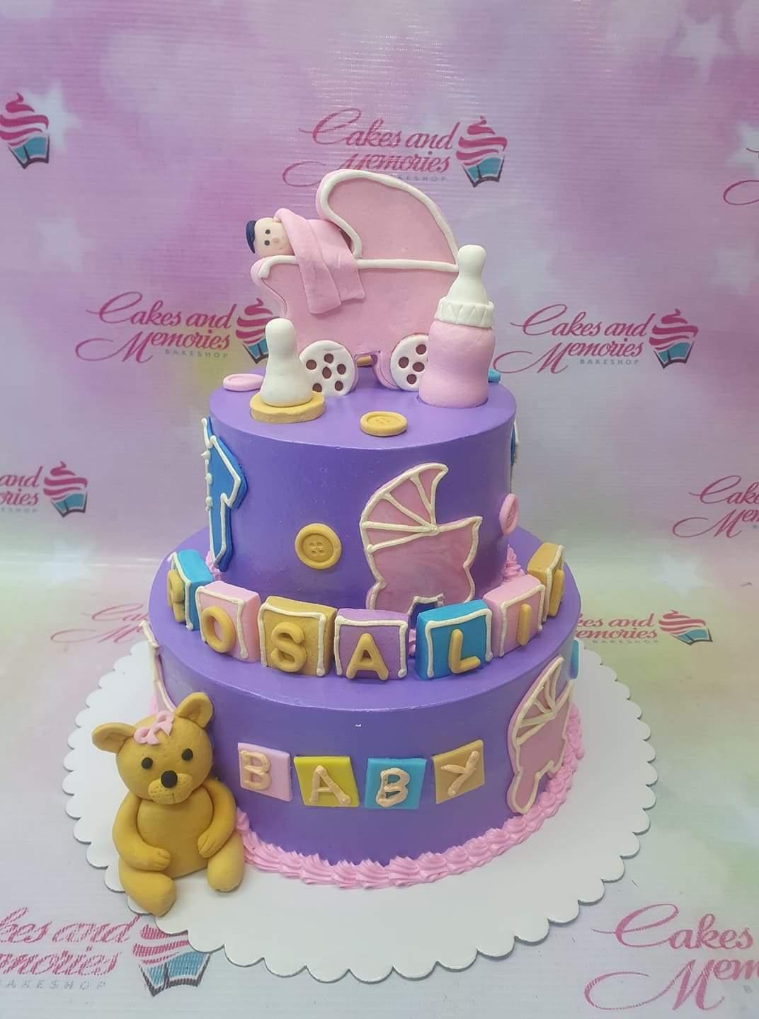 Two-tier purple fondant baby shower cake with a pink fondant baby stroller topper, a teddy bear, and colorful alphabet blocks.
