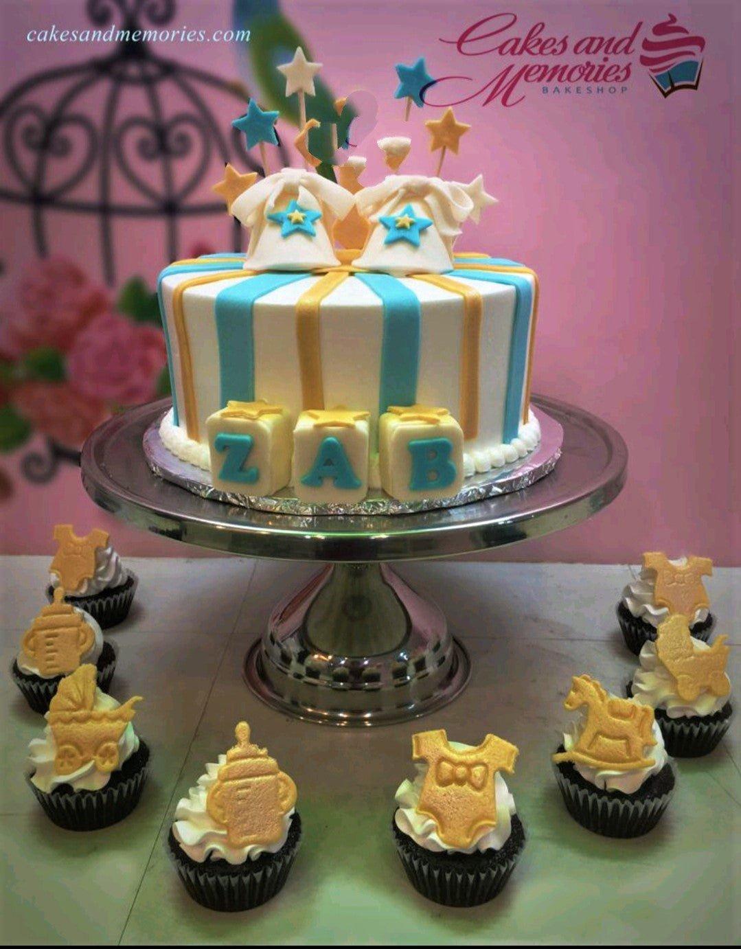 White fondant cake with blue and yellow vertical stripes, featuring baby clothes toppers and alphabet blocks for a baby shower.