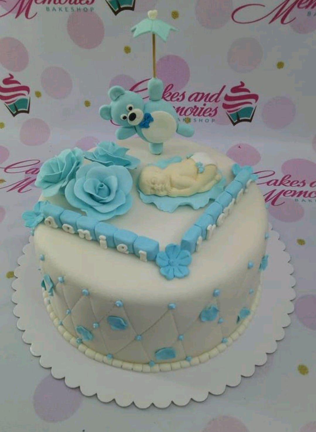White fondant baby shower cake with a blue teddy bear topper, sleeping baby figure, and blue roses.