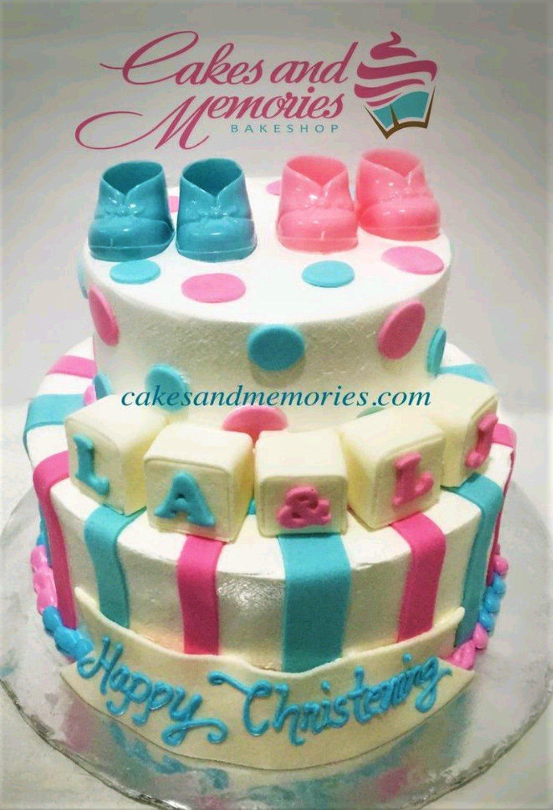 Two-tier baby shower cake with pink and blue stripes, polka dots, and fondant baby shoes on top.