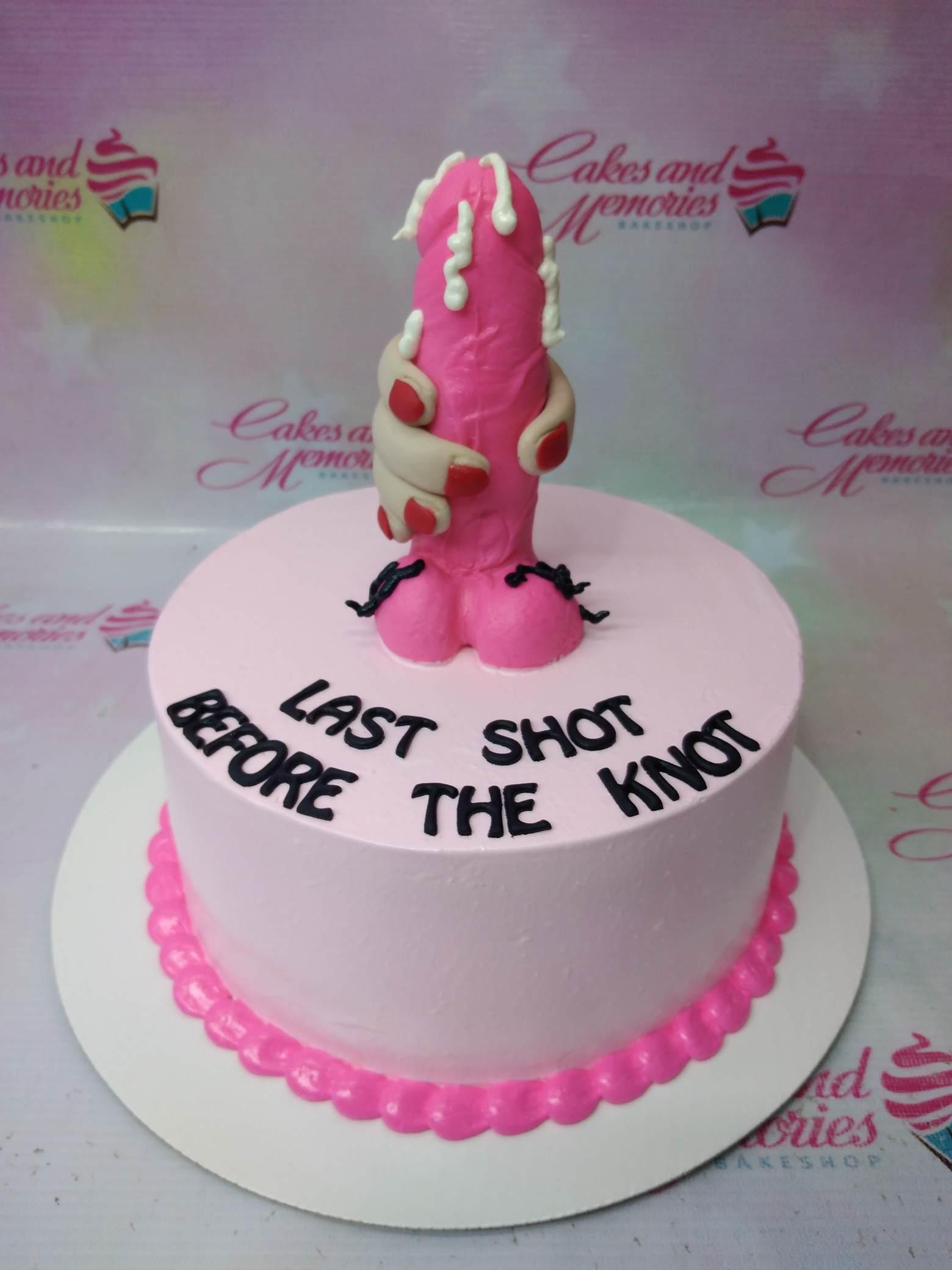 Pink bachelorette cake with a humorous pink phallic topper held by a hand and the message Last Shot Before The Knot.