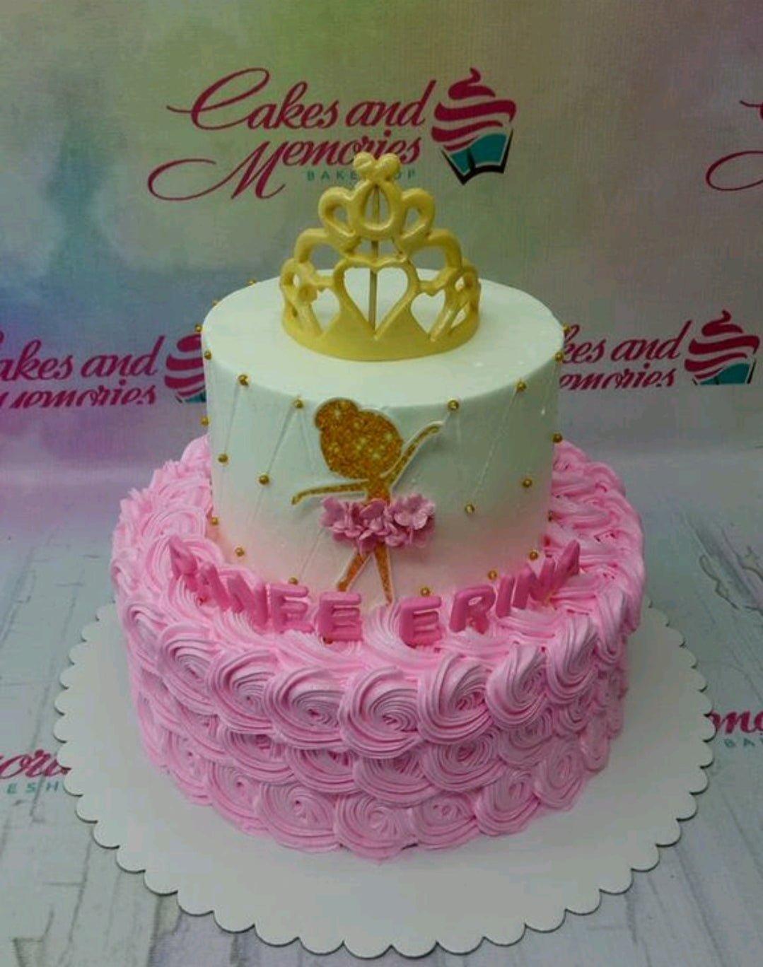 Two-tier pink and white ballerina birthday cake with a gold crown topper and pink icing rosettes.