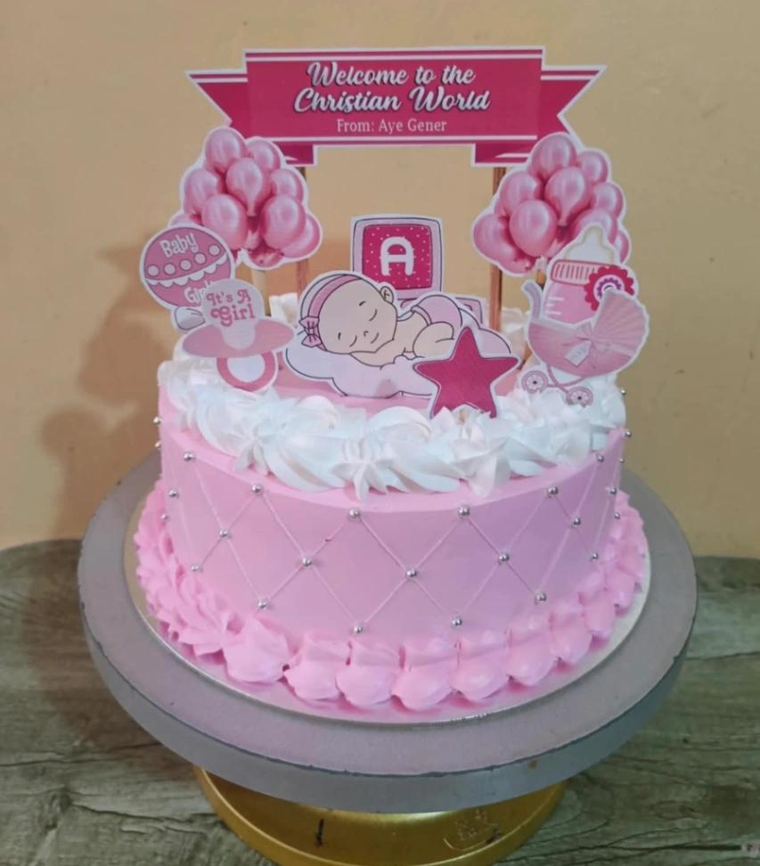 Pink Christian World baptism cake with a sleeping baby printout topper, pink balloons, and silver dragees on a quilted icing pattern.