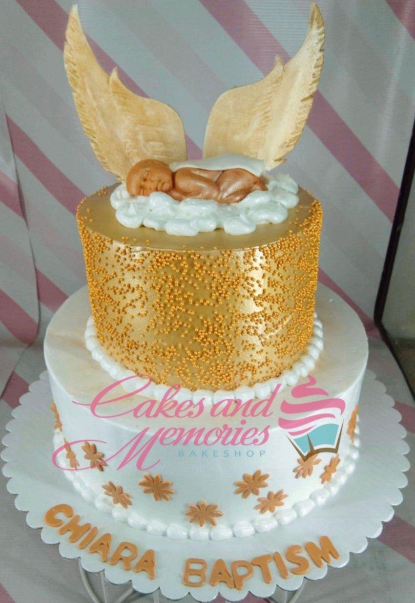 Two-tier gold and white baptism cake featuring a sleeping baby on clouds with large angel wings and gold sprinkles.
