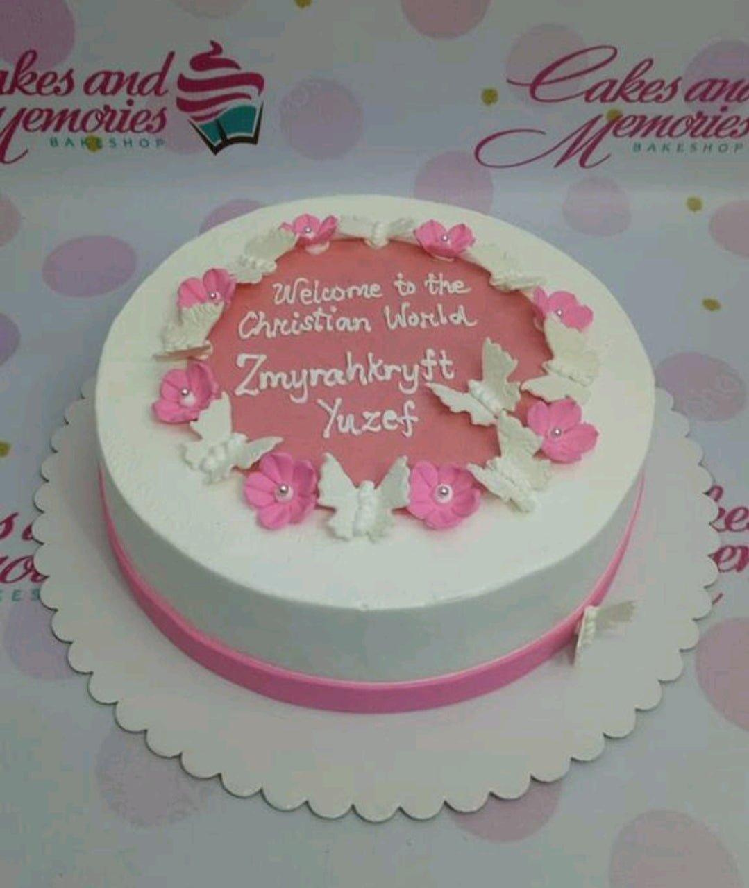 White and pink round cake for a Christian baptism celebration featuring white butterflies and pink flowers with a personalized message.