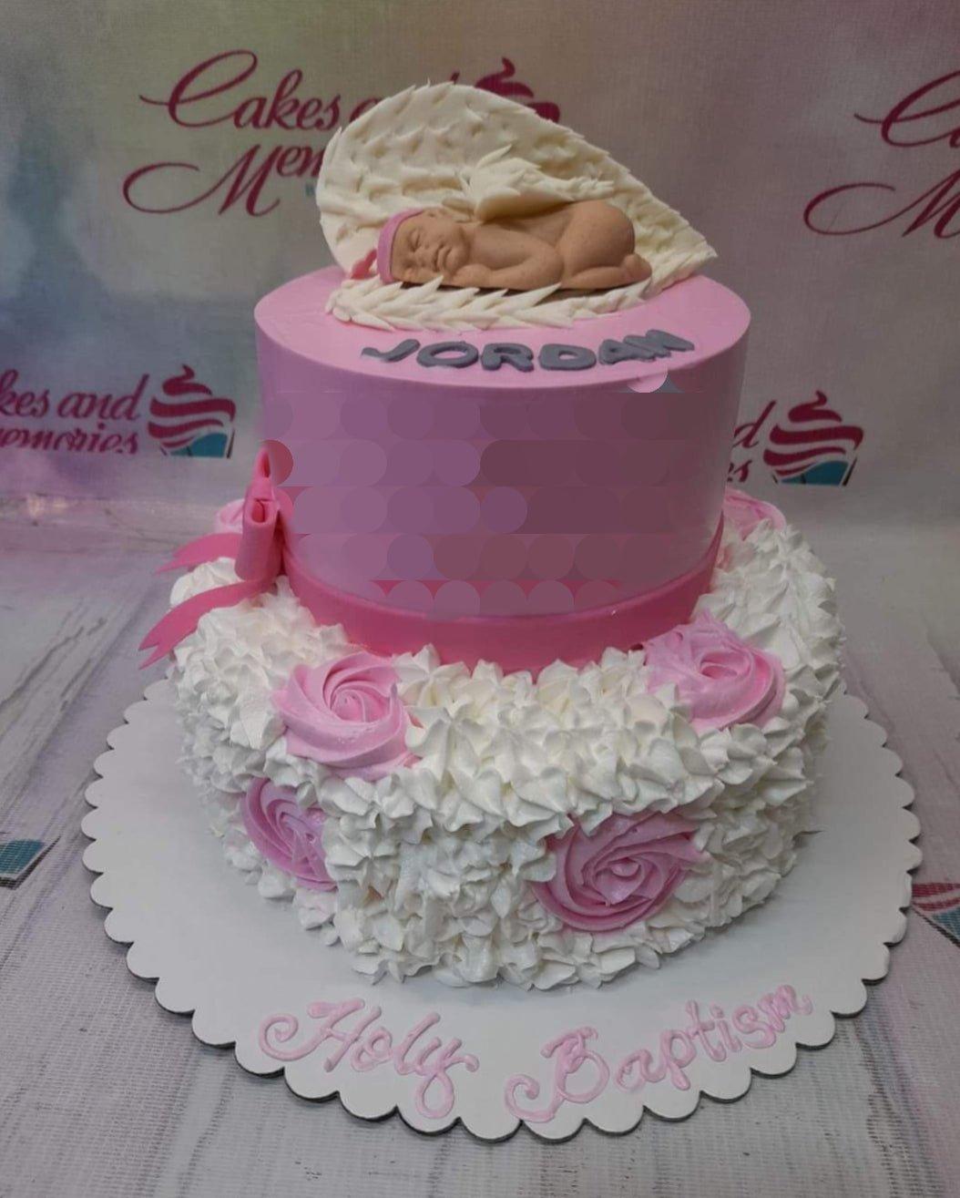 Pink and white 2-tier baptism cake with a sleeping baby angel topper and pink icing rosettes.