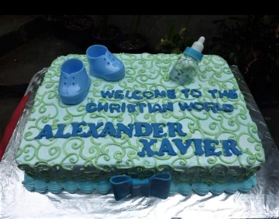 Light blue rectangular baptism cake with green icing swirls, blue baby shoes, a baby bottle topper, and a blue bow.