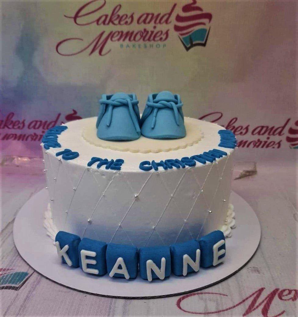 White round baptism cake with blue fondant baby shoes, quilted icing pattern with silver pearls, and blue block letters for Keane.