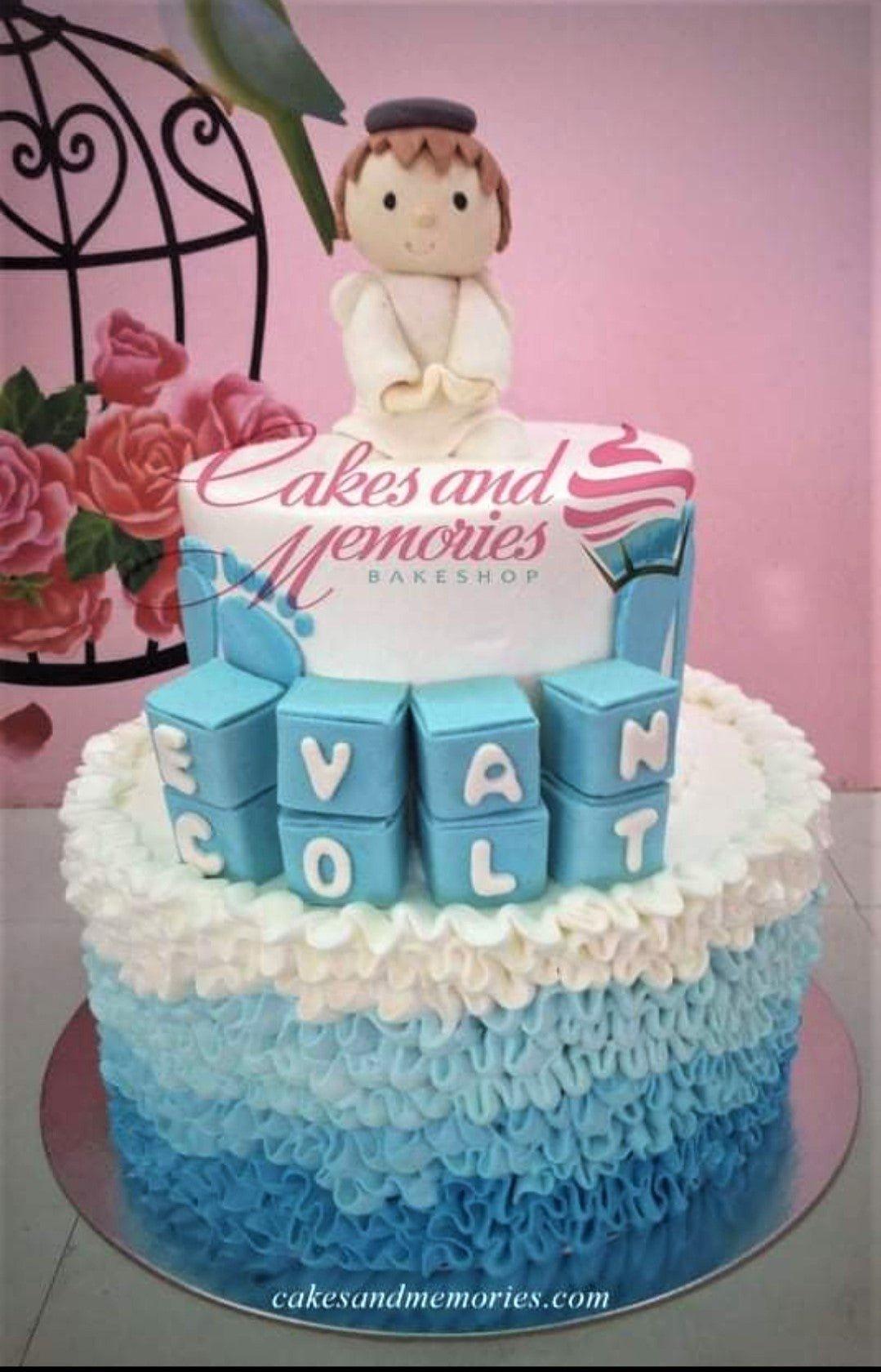 Two-tier blue and white baptism cake with a fondant angel topper, blue blocks spelling EVAN COLT, and ombre blue icing ruffles.