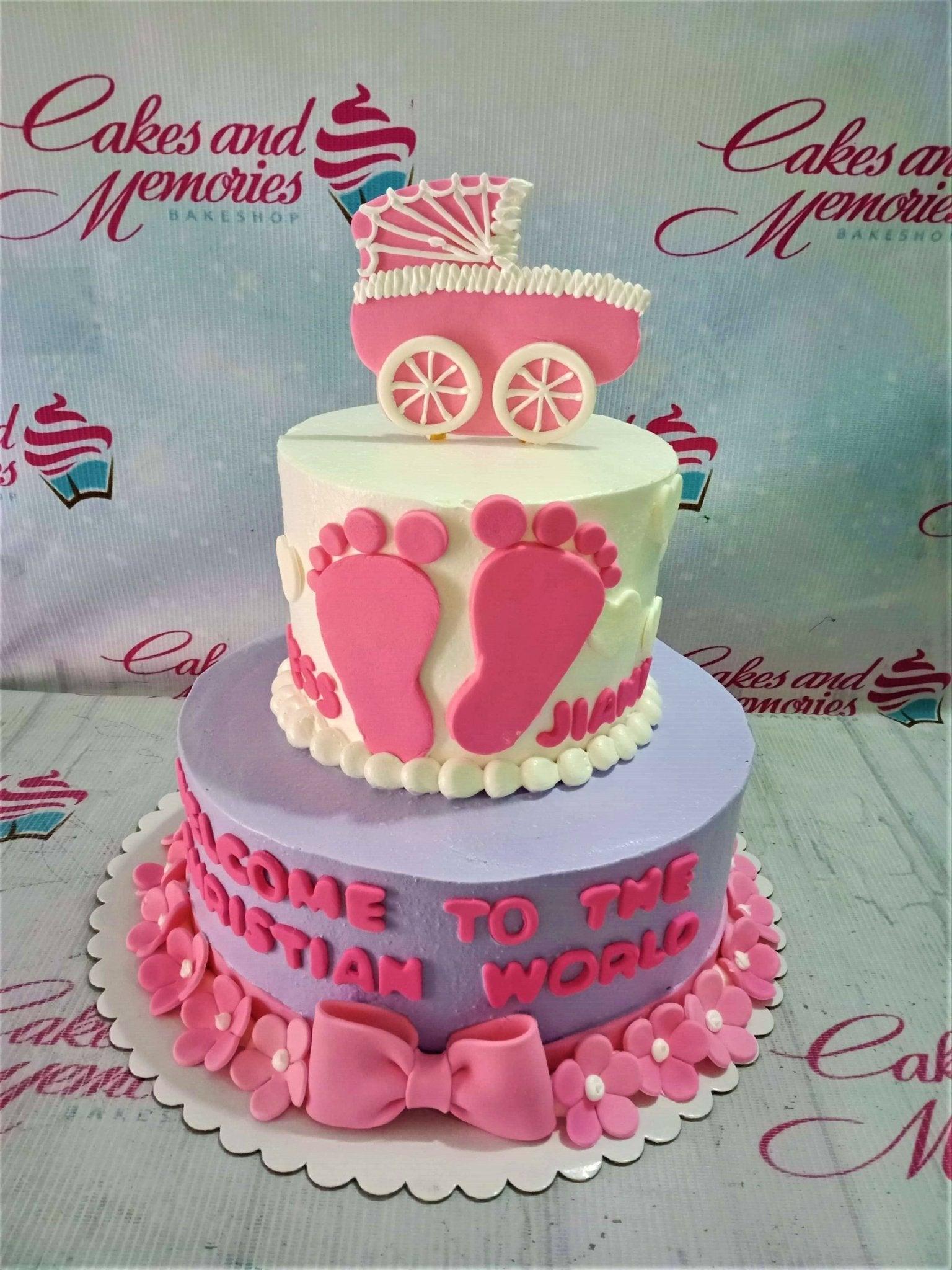 Two-tier baby girl baptism cake with pink baby carriage topper, pink footprints, and floral base border in purple and white icing.