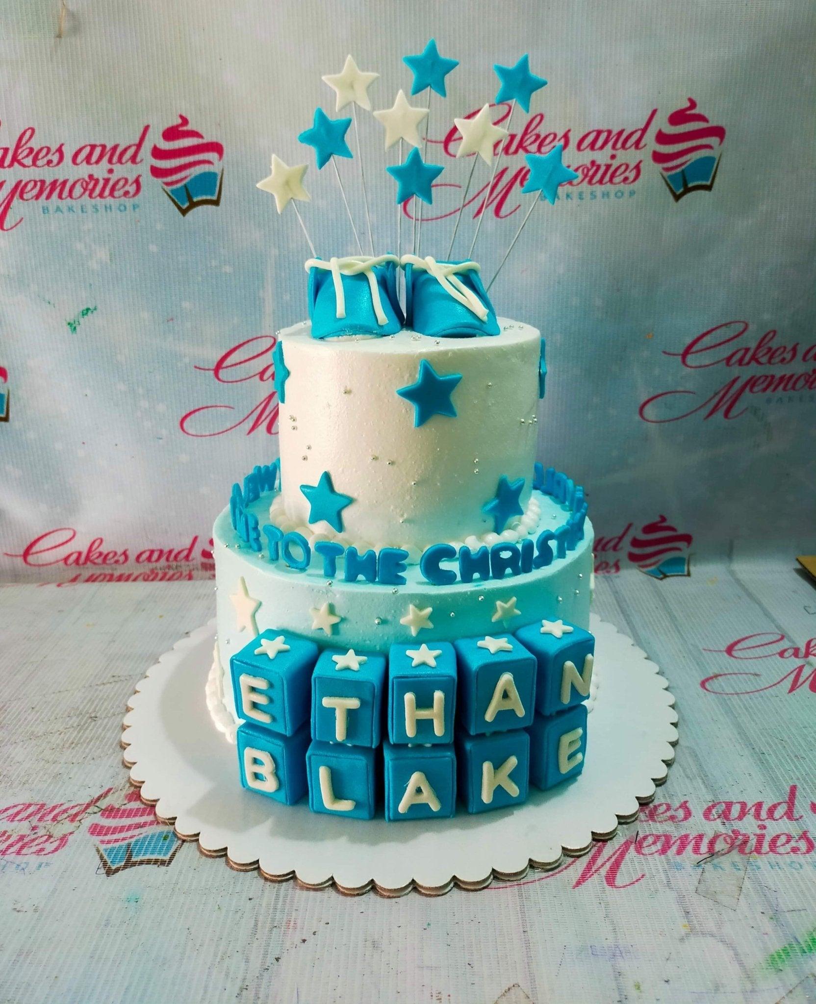 Two-tier blue and white baby boy baptism cake with fondant baby shoes, stars, and name blocks.
