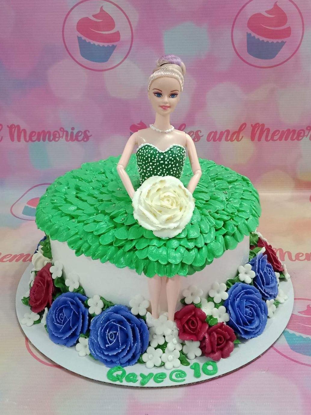 Barbie doll cake with a green ruffled icing dress and a white rose, surrounded by blue and red icing flowers on a white base.