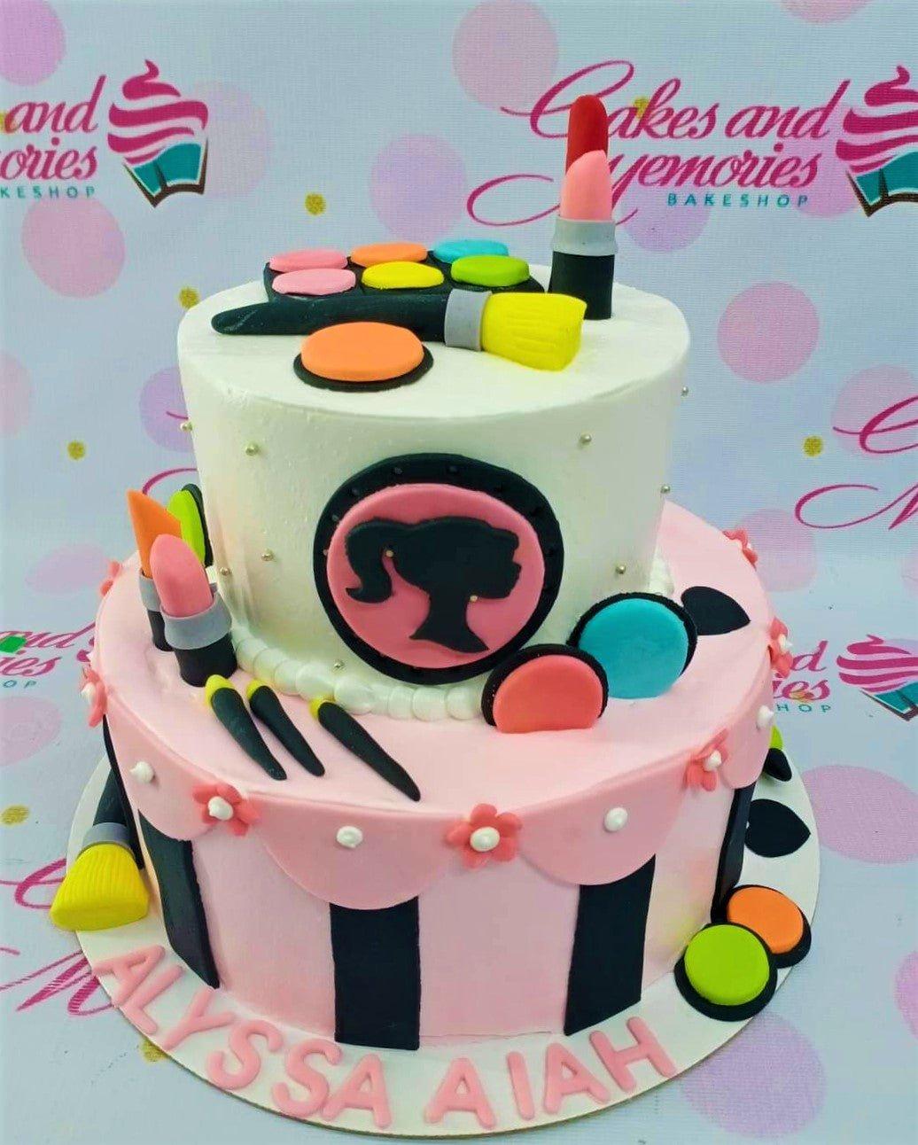 Two-tier pink and white Barbie makeup themed cake with fondant lipstick, eyeshadow palette, and brushes.
