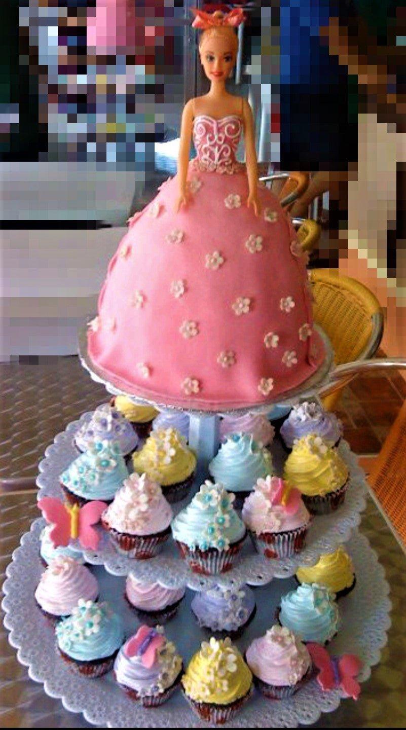 Barbie doll cake with a pink fondant ball gown decorated with white flowers, displayed on a tiered stand with colorful cupcakes.