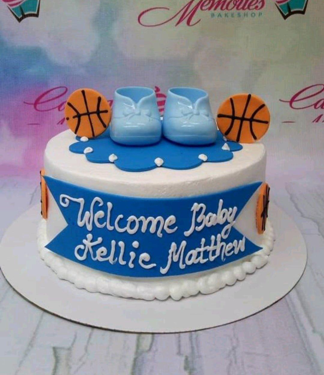 White 1-tier basketball and baby shoes themed cake with blue accents and a personalized message banner.