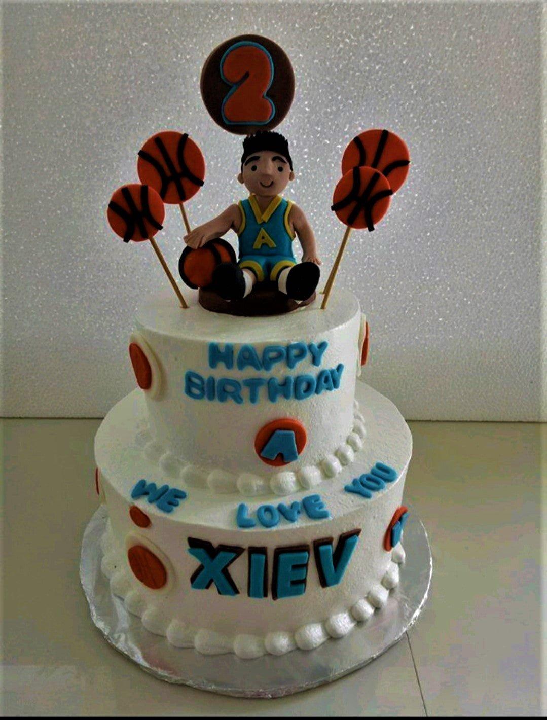 Two-tier basketball themed birthday cake with a 3D basketball player figure, basketball toppers, and blue icing text.