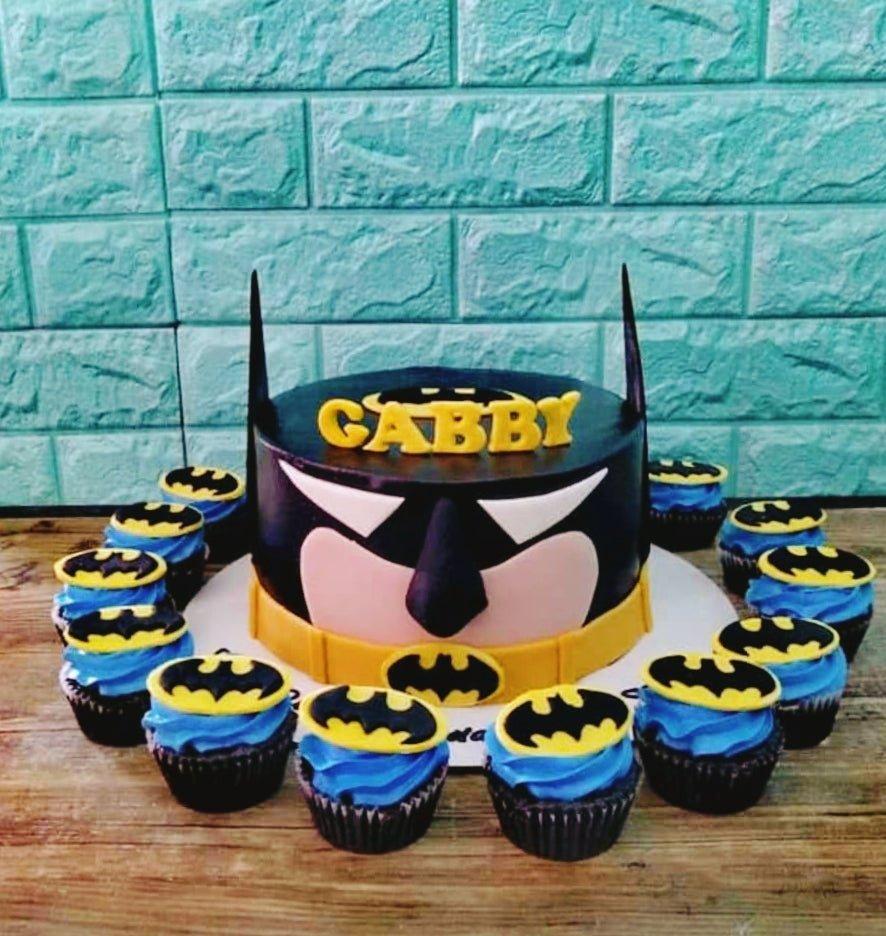 Batman themed birthday cake with black icing, yellow Batman logo belt, and matching blue cupcakes with Batman toppers.