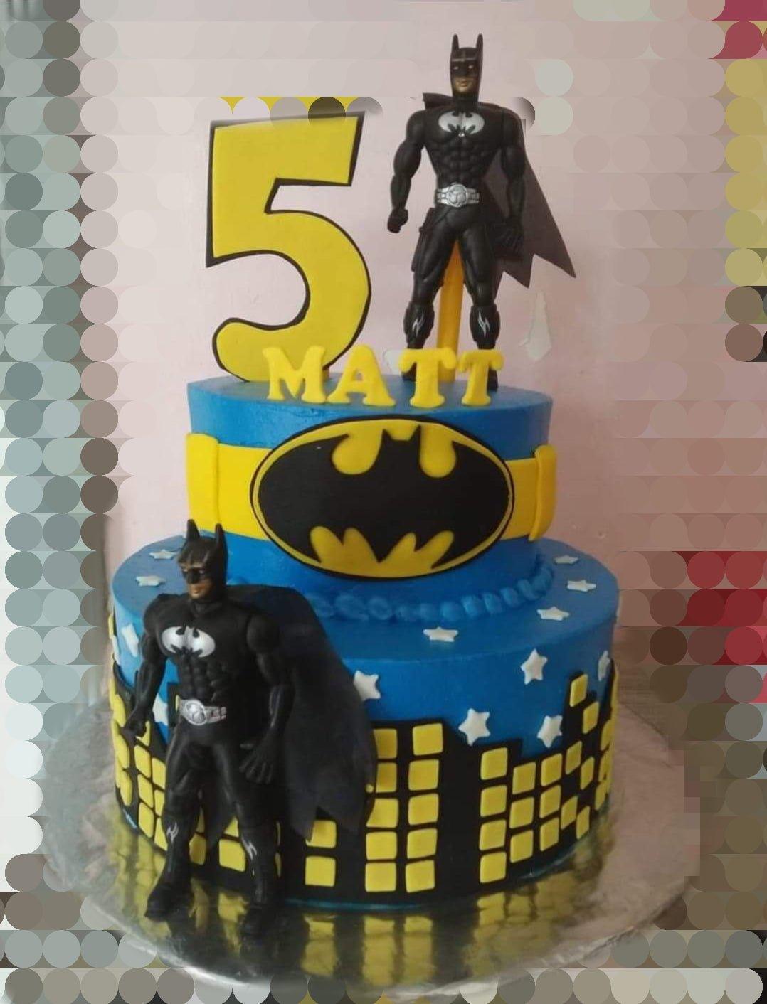 Two-tier blue Batman themed birthday cake with city skyline, stars, and Batman figurines.