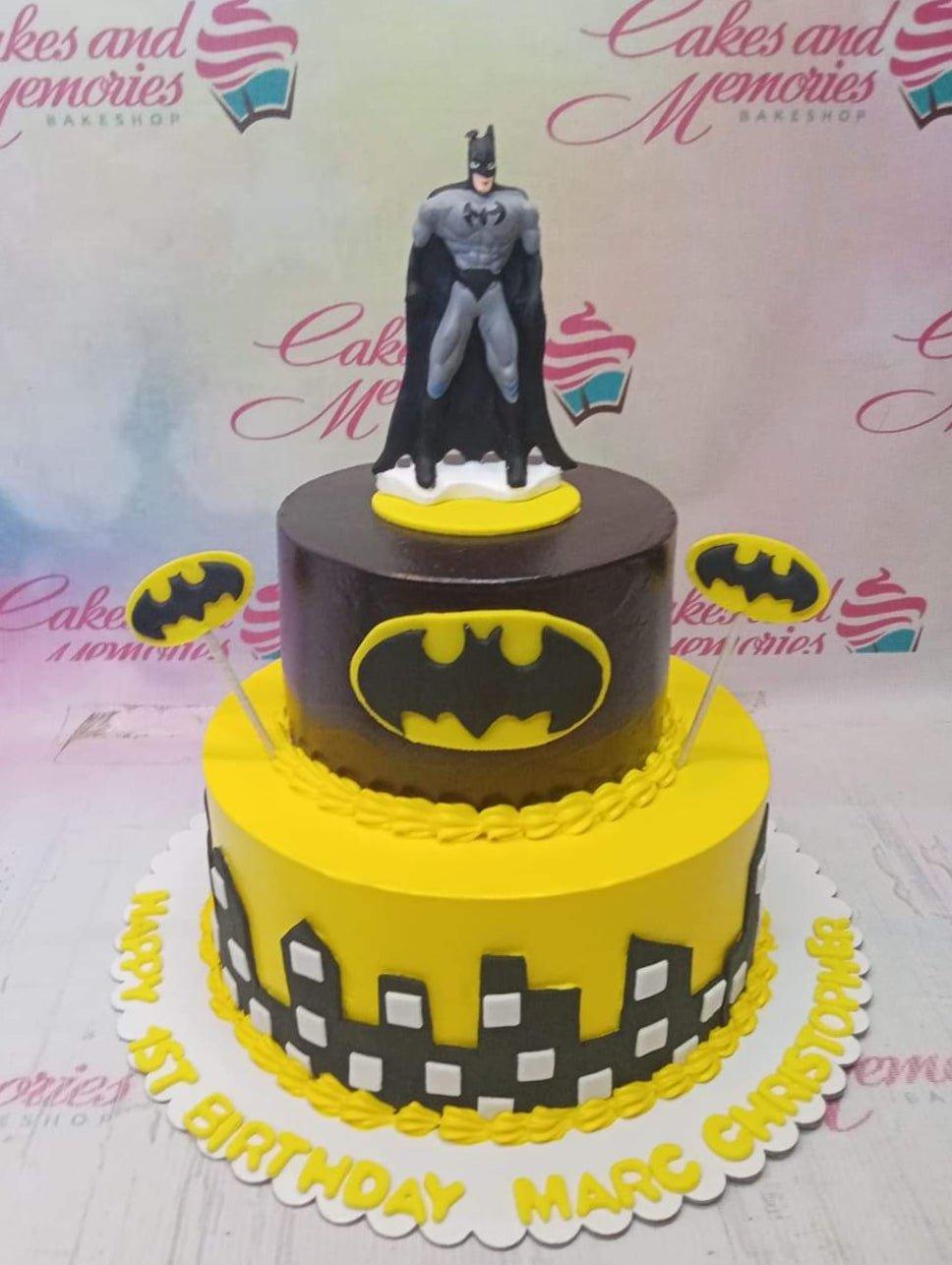 Two-tier Batman themed birthday cake in yellow and black with a Batman figurine topper and city skyline silhouette.