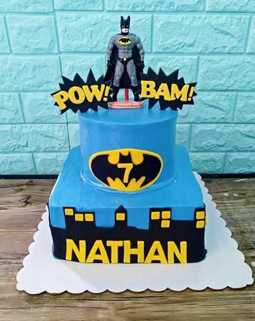 Two-tier Batman themed birthday cake in light blue with a Batman toy figure, city skyline silhouette, and yellow name letters.