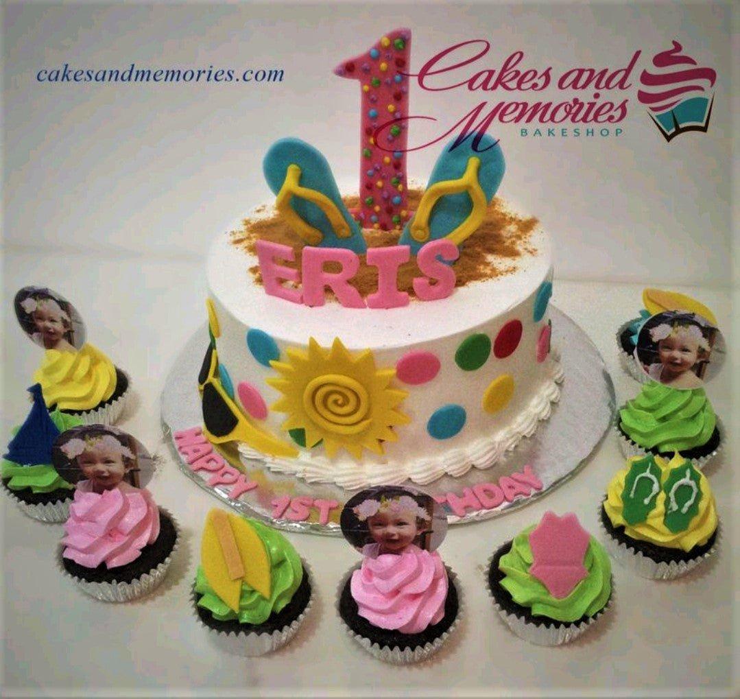 Beach themed birthday cake with pink fondant name Eris, flip flops, sun, and surfboard decorations surrounded by cupcakes.