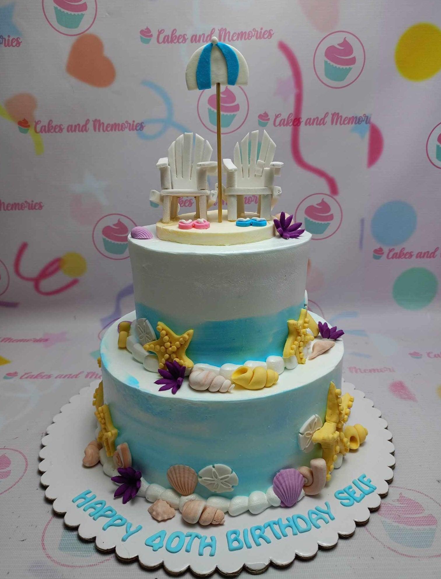 Two-tier beach themed birthday cake with blue and white gradient icing, fondant beach chairs, umbrella, and various seashells.