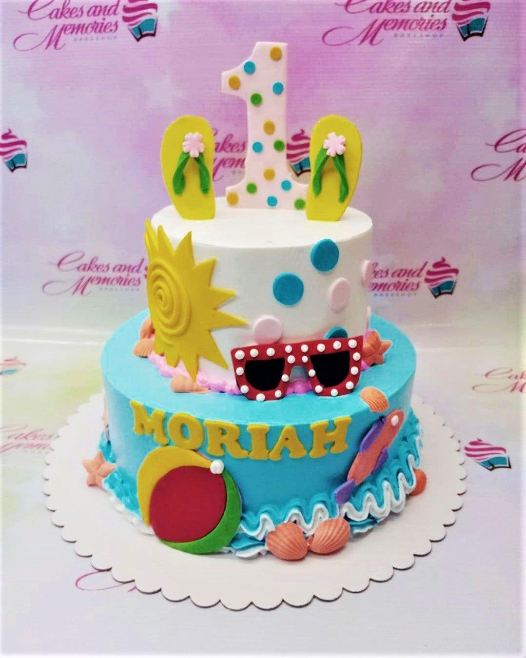 Two-tier beach themed birthday cake with a white top tier and blue bottom tier featuring a sun, sunglasses, flip flops, and a beach ball.