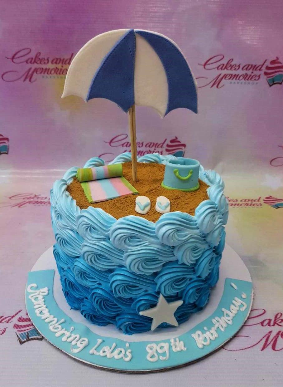 Beach themed birthday cake with blue icing waves, sand top, beach umbrella, and a blue bucket topper.
