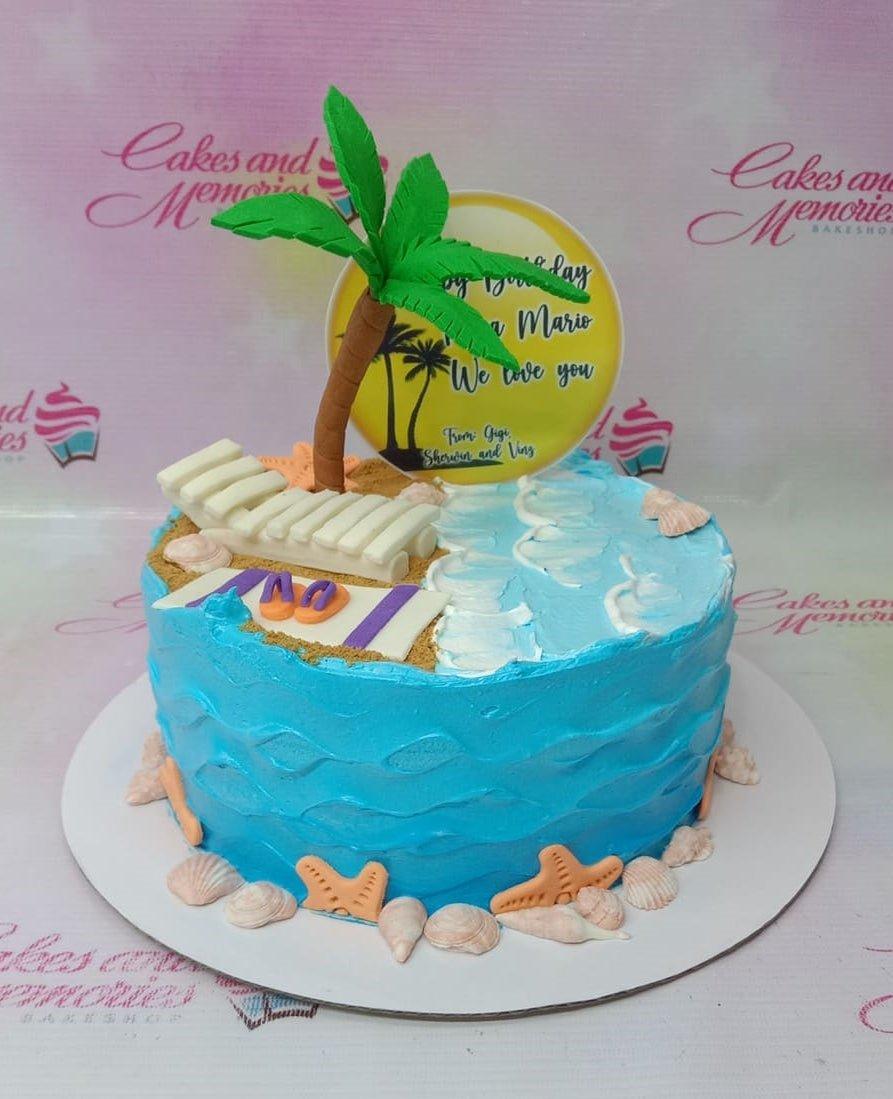 Blue beach themed birthday cake with a fondant palm tree, beach chair, and edible seashells on a sandy base.