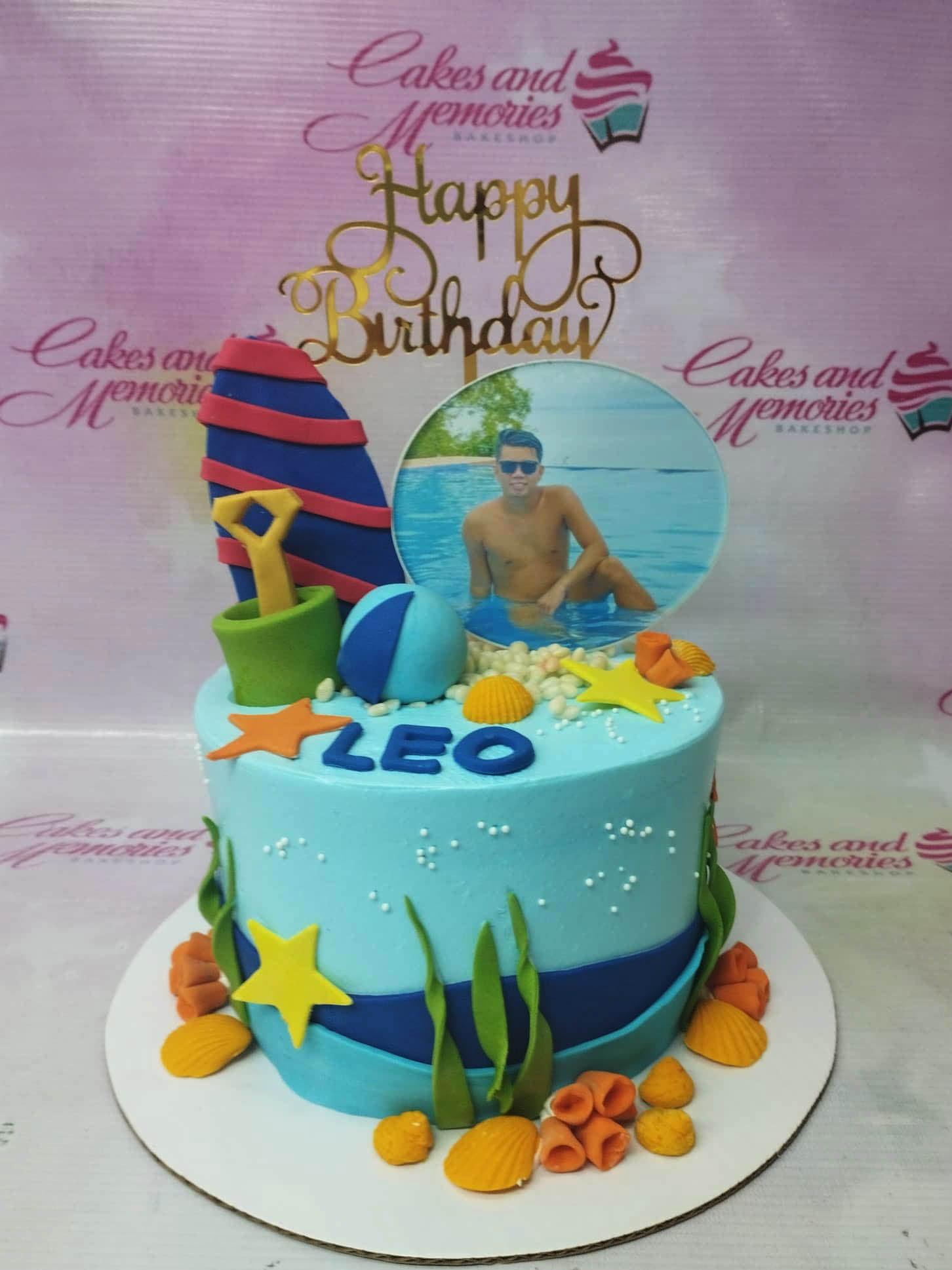 Light blue beach themed cake with a surfboard, beach ball, sand bucket, and a photo printout of a man in a pool.
