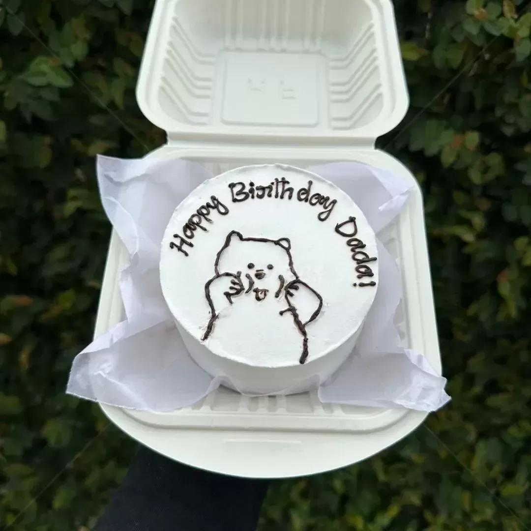 White bento cake in a clamshell container featuring a hand-drawn black icing doodle of a cute bear and 'Happy Birthday Dada!' message.