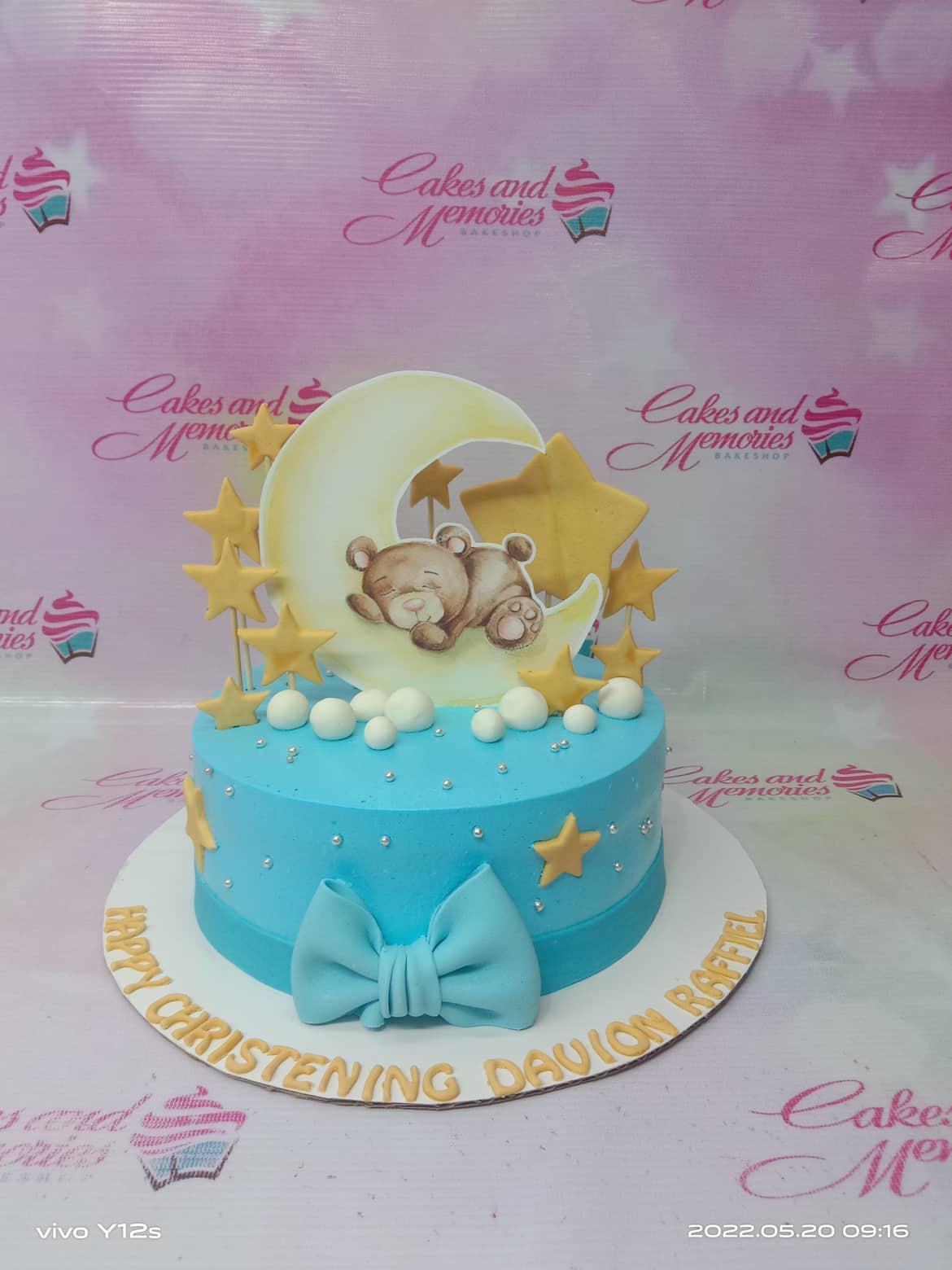 Light blue christening cake with a sleeping bear on a crescent moon topper, yellow stars, and a large blue bow.