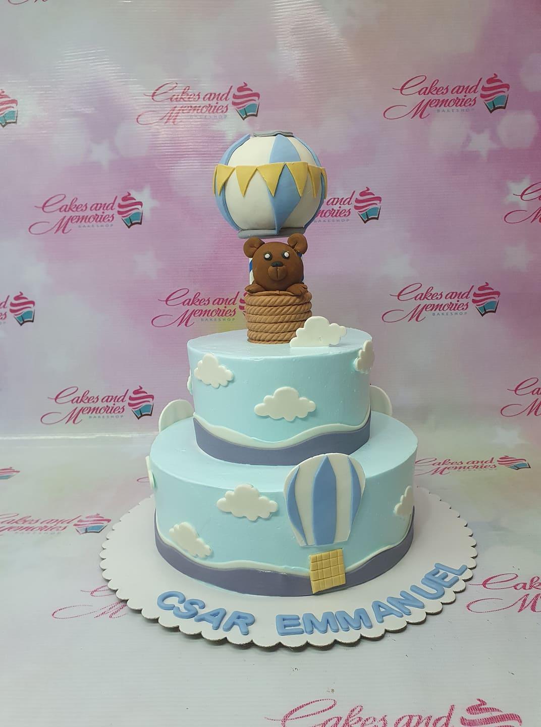 Two-tier light blue fondant cake with a 3D bear in a hot air balloon topper, white clouds, and blue gumpaste letters on the base.