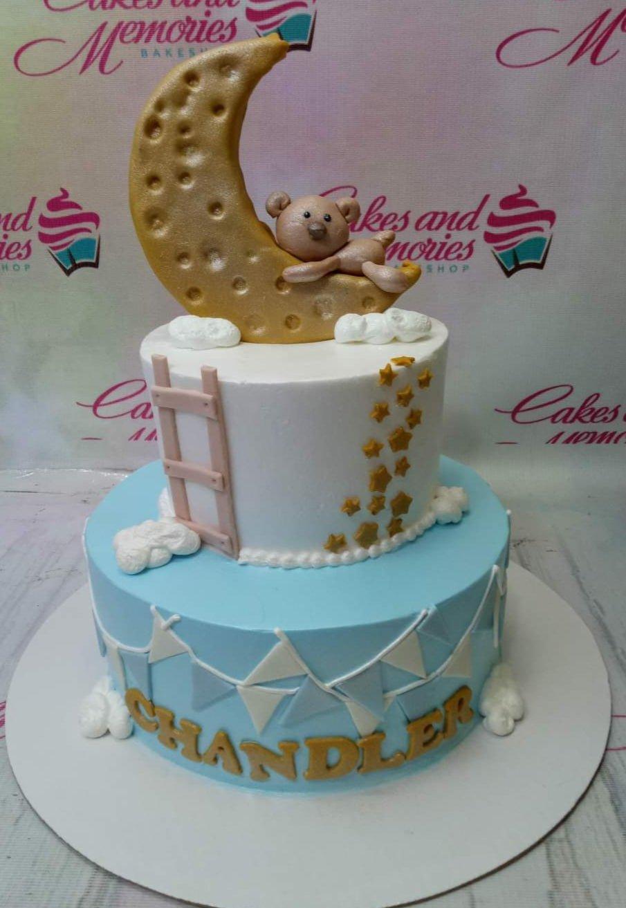 Two-tier blue and white baby shower cake with a fondant bear sleeping on a gold moon topper, stars, and a ladder.