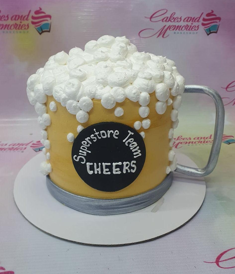 Beer mug cake with yellow icing, white foam dollops, a silver handle, and a black circular message plaque.