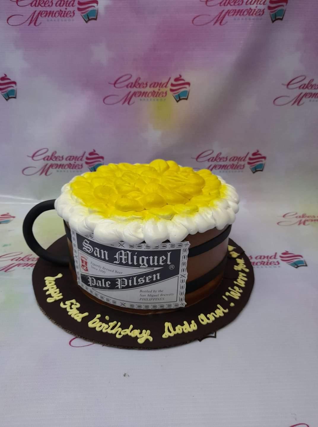 San Miguel Pale Pilsen beer mug cake with yellow and white icing foam and a black handle on a brown base board.