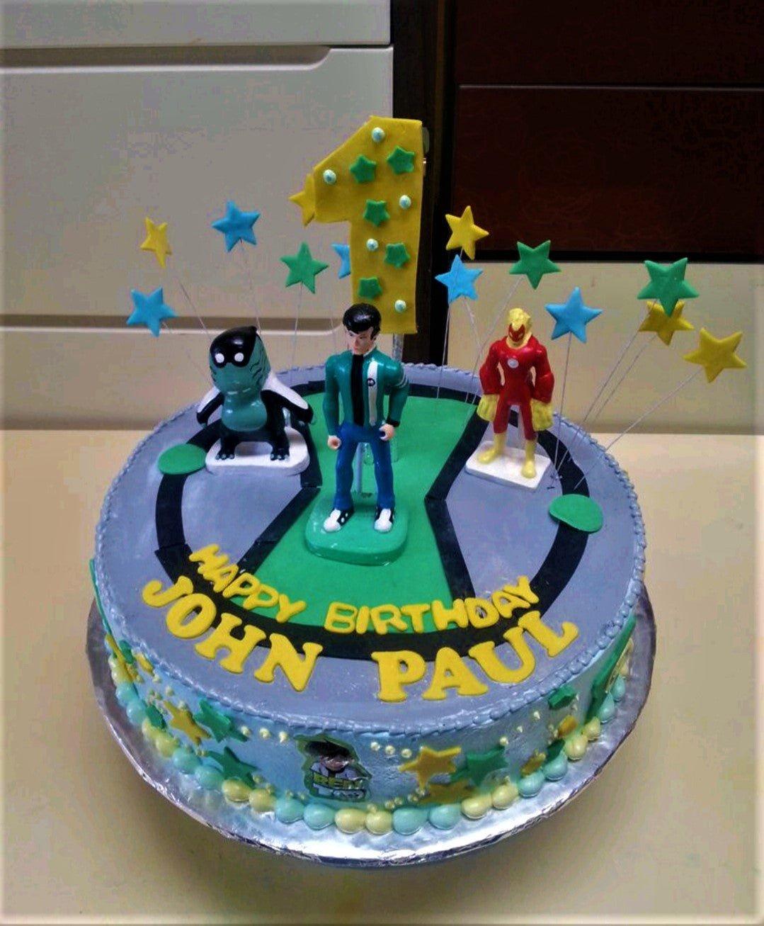 Ben 10 themed birthday cake with character toys, green and black Omnitrix design, and yellow gumpaste letters for John Paul.