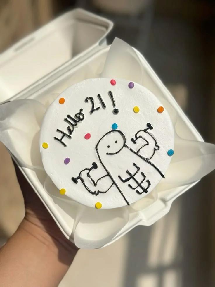 White bento cake with a hand-drawn black icing doodle of a character lifting weights and a sewing machine, accented with colorful dots.
