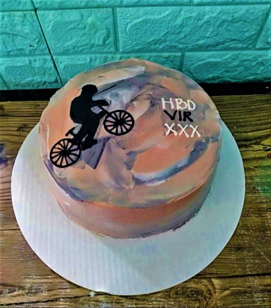 Bento cake with a textured orange and purple palette knife icing design featuring a black bicycle rider silhouette and a birthday message.