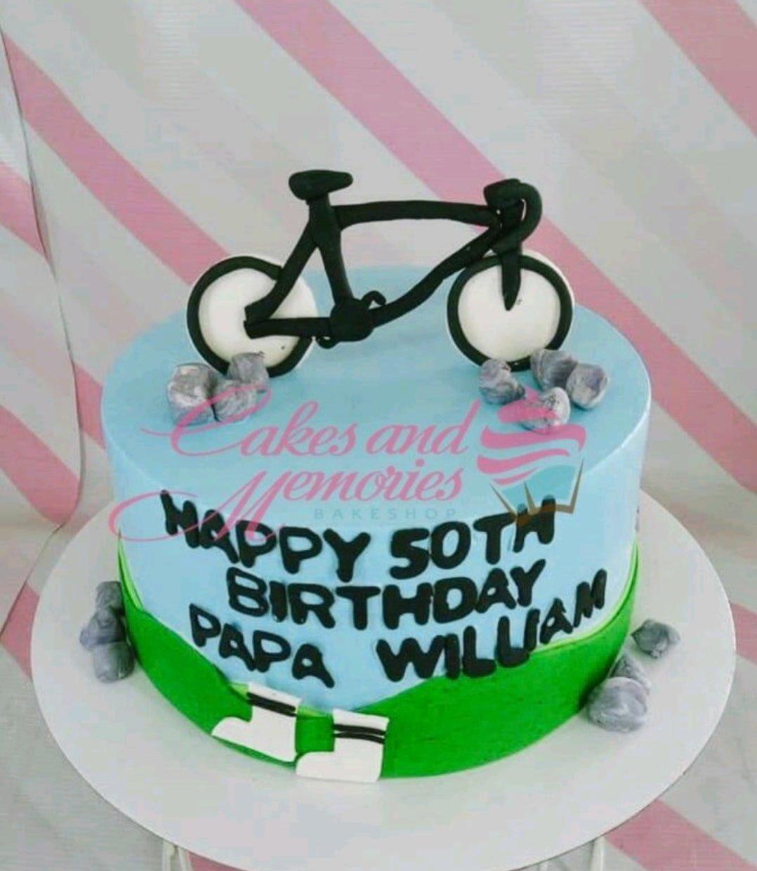 Light blue 50th birthday cake for Papa William featuring a black bicycle topper, fondant rocks, and green grass icing details.