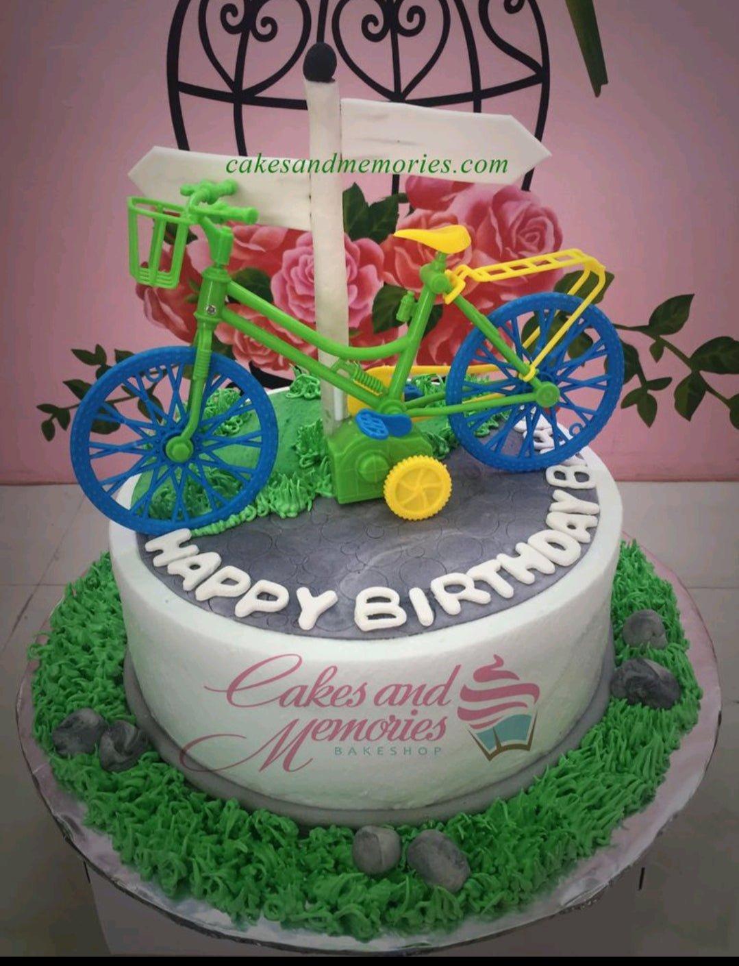 A bicycle themed birthday cake with a green plastic toy bike, white signpost, and grey road icing design.