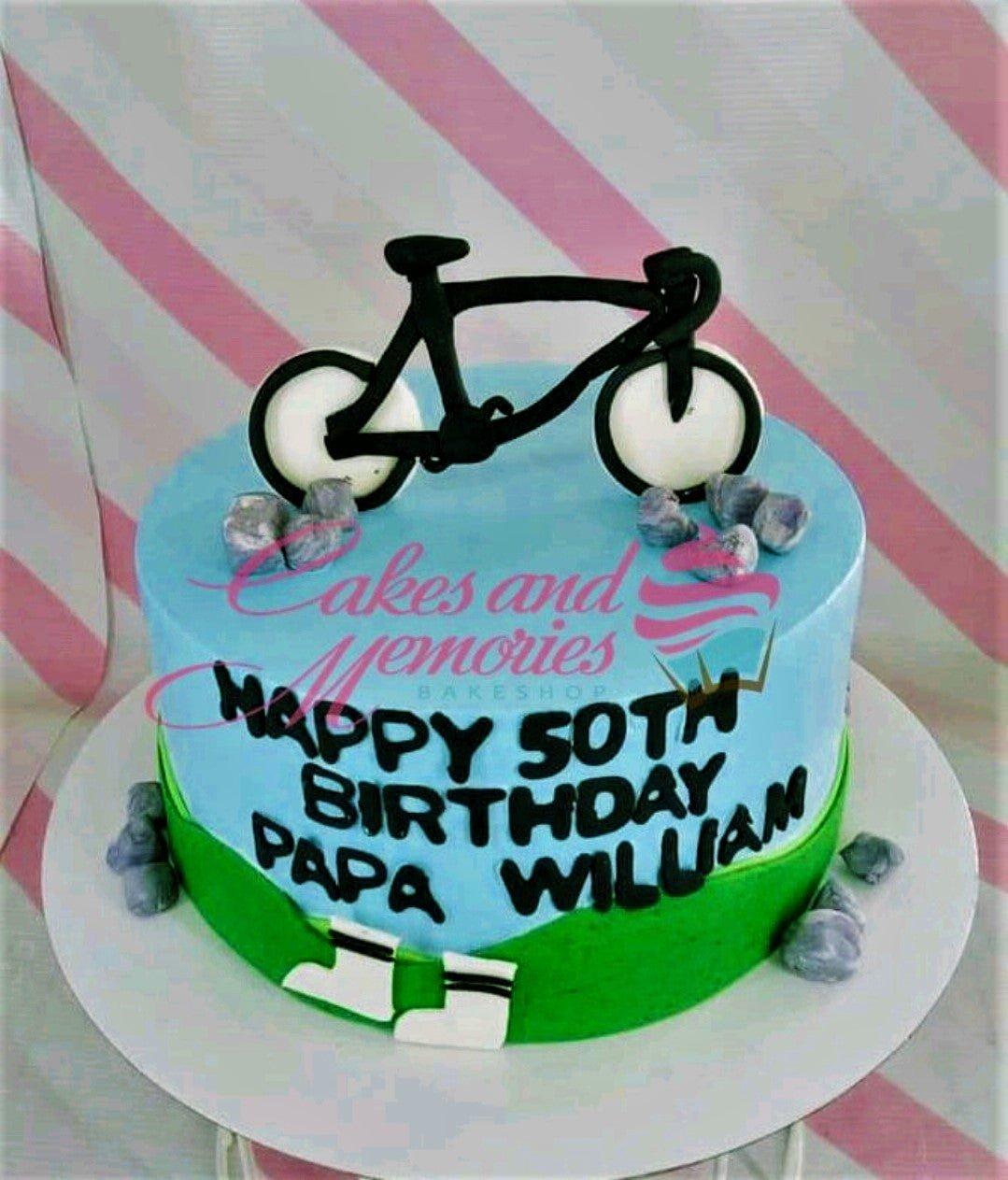 Light blue 1-tier bicycle themed birthday cake with a black bicycle topper, fondant rocks, and green grass icing at the base.