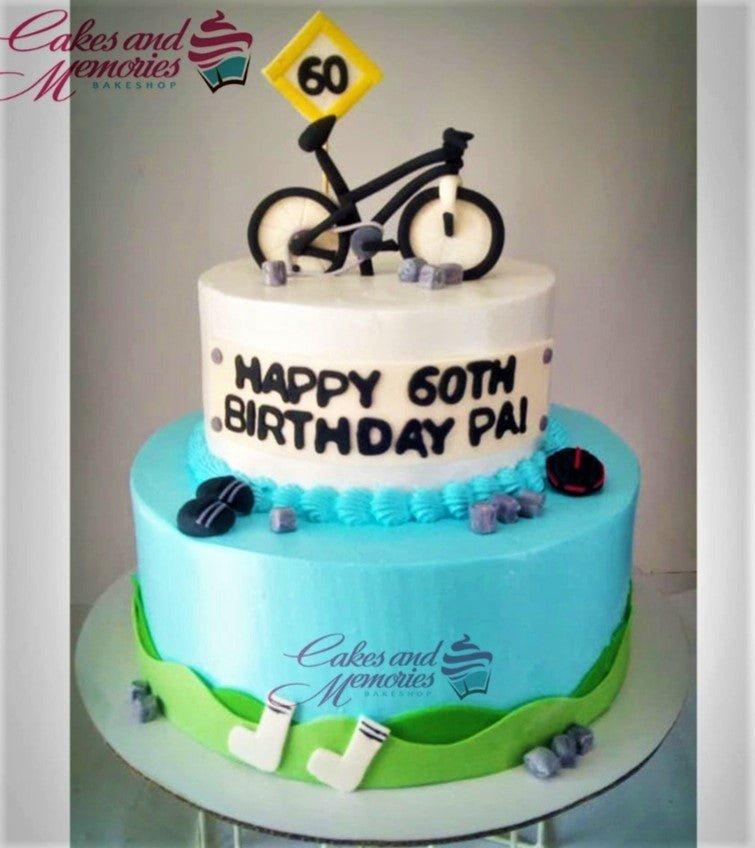 Two-tier bicycle themed birthday cake with light blue and white icing, featuring a 3D bicycle topper and green grass accents.