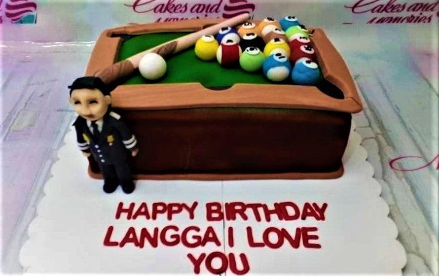 Billiard table cake with fondant pool balls, cue stick, and a pilot figurine on a white board with a birthday message.