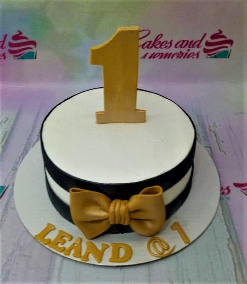 A white fondant cake with black horizontal stripes, a large gold fondant bow, and a gold number one topper.