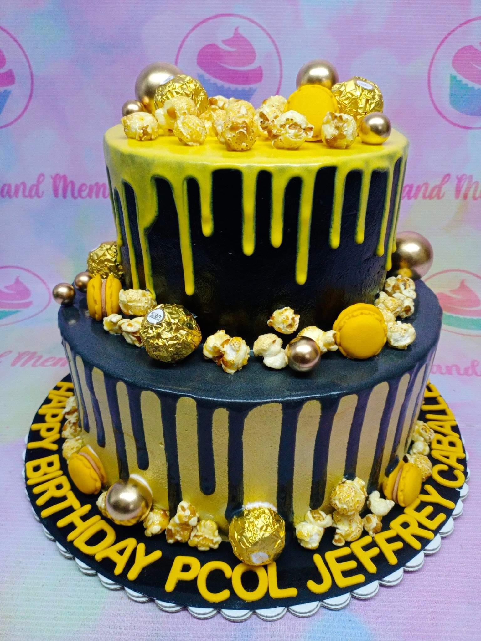 Black and gold 2-tier birthday cake with yellow chocolate drip, Ferrero Rocher, popcorn, and yellow macarons.