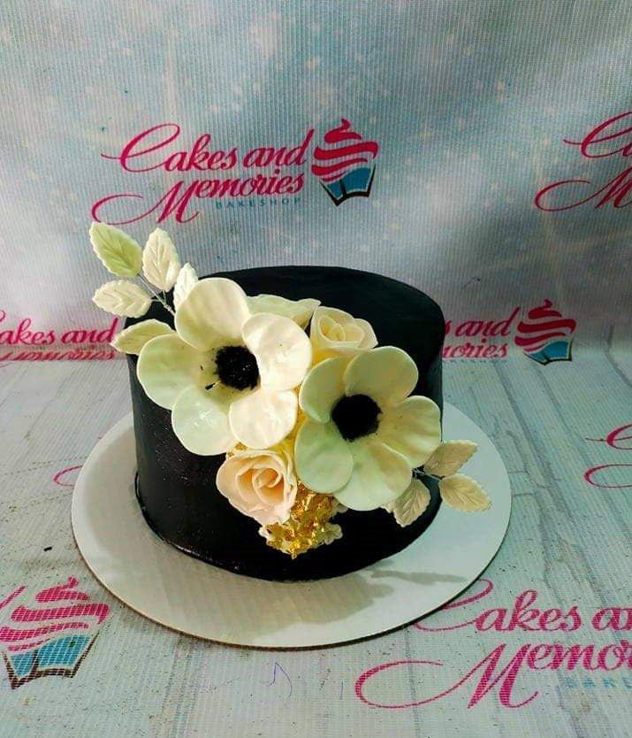 Elegant black 1-tier cake decorated with large white gumpaste flowers and gold leaf accents.