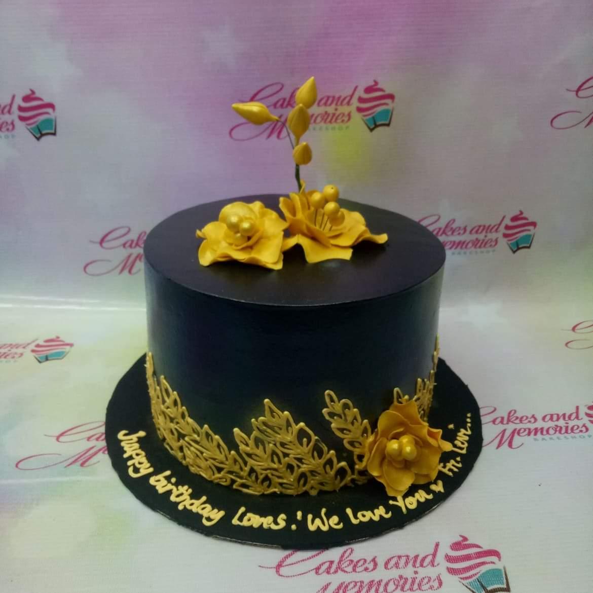 Elegant black 1-tier cake with gold gumpaste flowers and intricate gold leaf patterns on the side.
