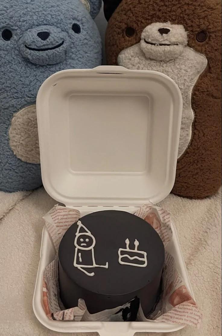 Black minimalist bento cake with white icing doodle of a person in a party hat sitting next to a birthday cake.